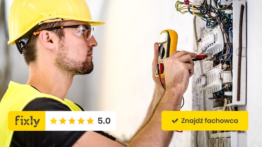 Elektryk w żółtym kasku i kamizelce ochronnej, z profilu, mierzy napięcie w skrzynce rozdzielczej za pomocą multimetru, widoczne przewody i bezpieczniki.