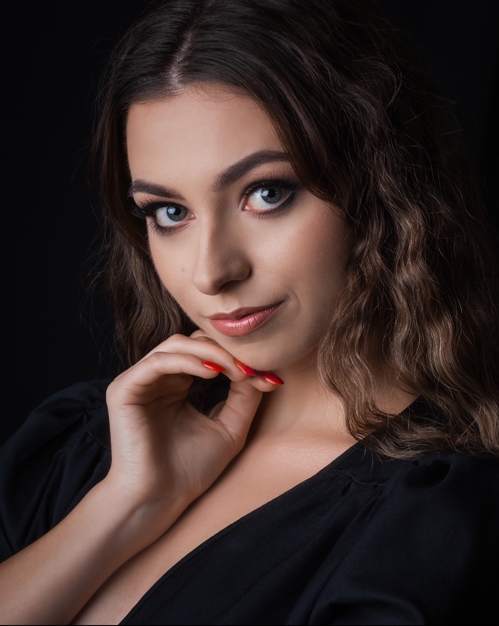 Portret kobiety z ciemnymi, kręconymi włosami, opierającej podbródek na dłoni z czerwonymi paznokciami, na czarnym tle.