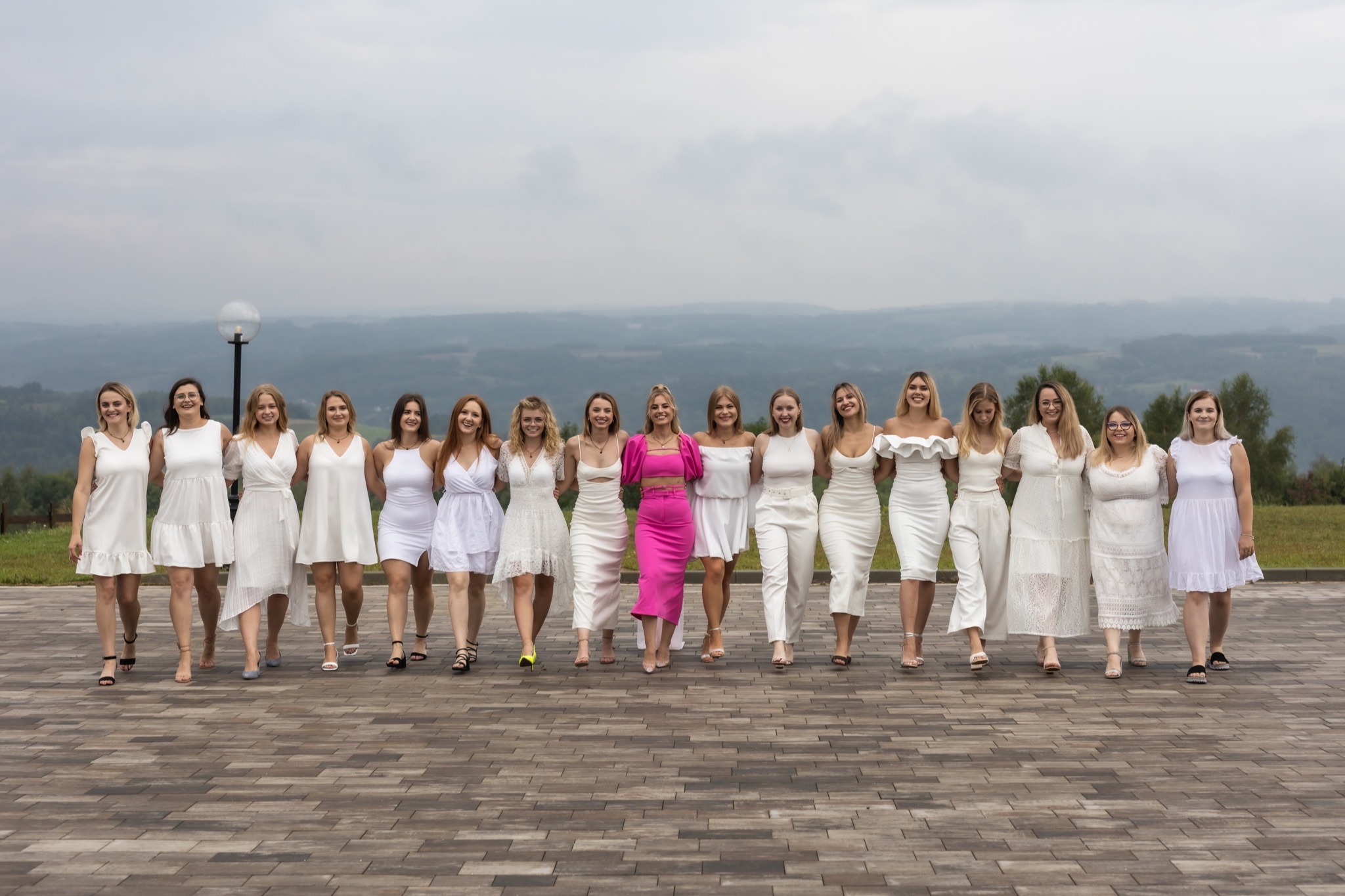 Grupowe zdjęcie trzynastu kobiet w białych sukienkach i butach na obcasie, idących po brukowanej ścieżce na tle mglistego krajobrazu wzgórz i pochmurnego nieba, z jedną kobietą ubraną w różowy...