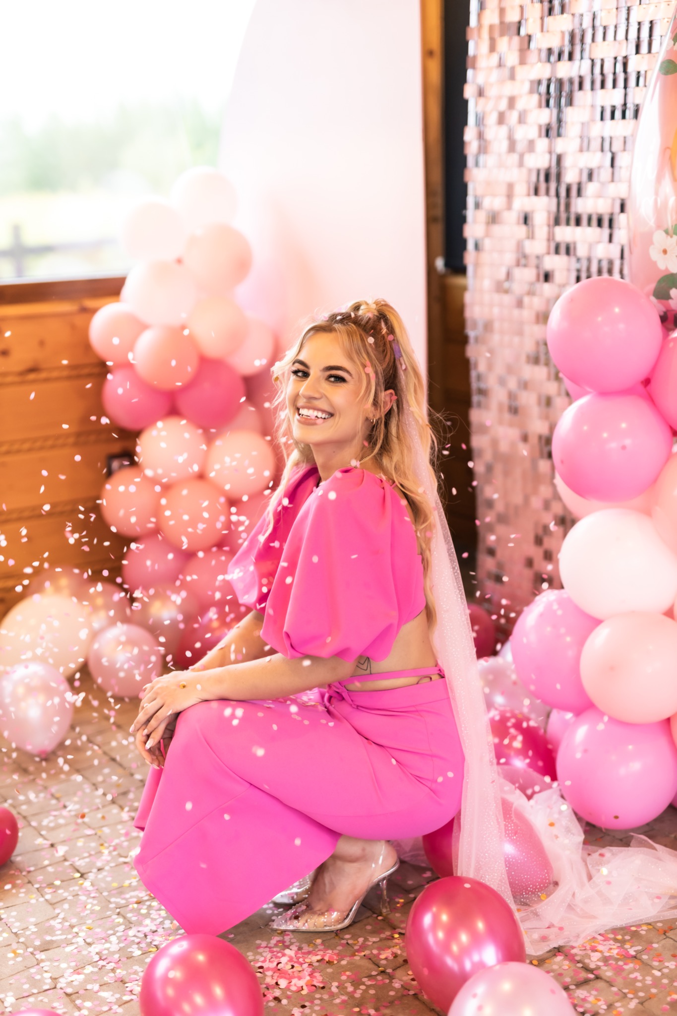 Uśmiechnięta blondynka w różowym kombinezonie pozuje w kucki wśród różowych balonów i konfetti, z przezroczystymi szpilkami na nogach i tiulowym welonem.