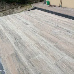 Ułożona kostka brukowa imitująca drewno na tarasie przydomowym, widoczne obrzeża z ciemnej kostki, w tle ziemia, trawa, konewka i wąż ogrodowy.