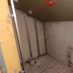 Wnętrze w trakcie remontu: widoczne metalowe profile ścian działowych, instalacja ogrzewania podłogowego na podłodze, surowy sufit z pomarańczowymi elementami mocującymi oraz wiadro z masą...