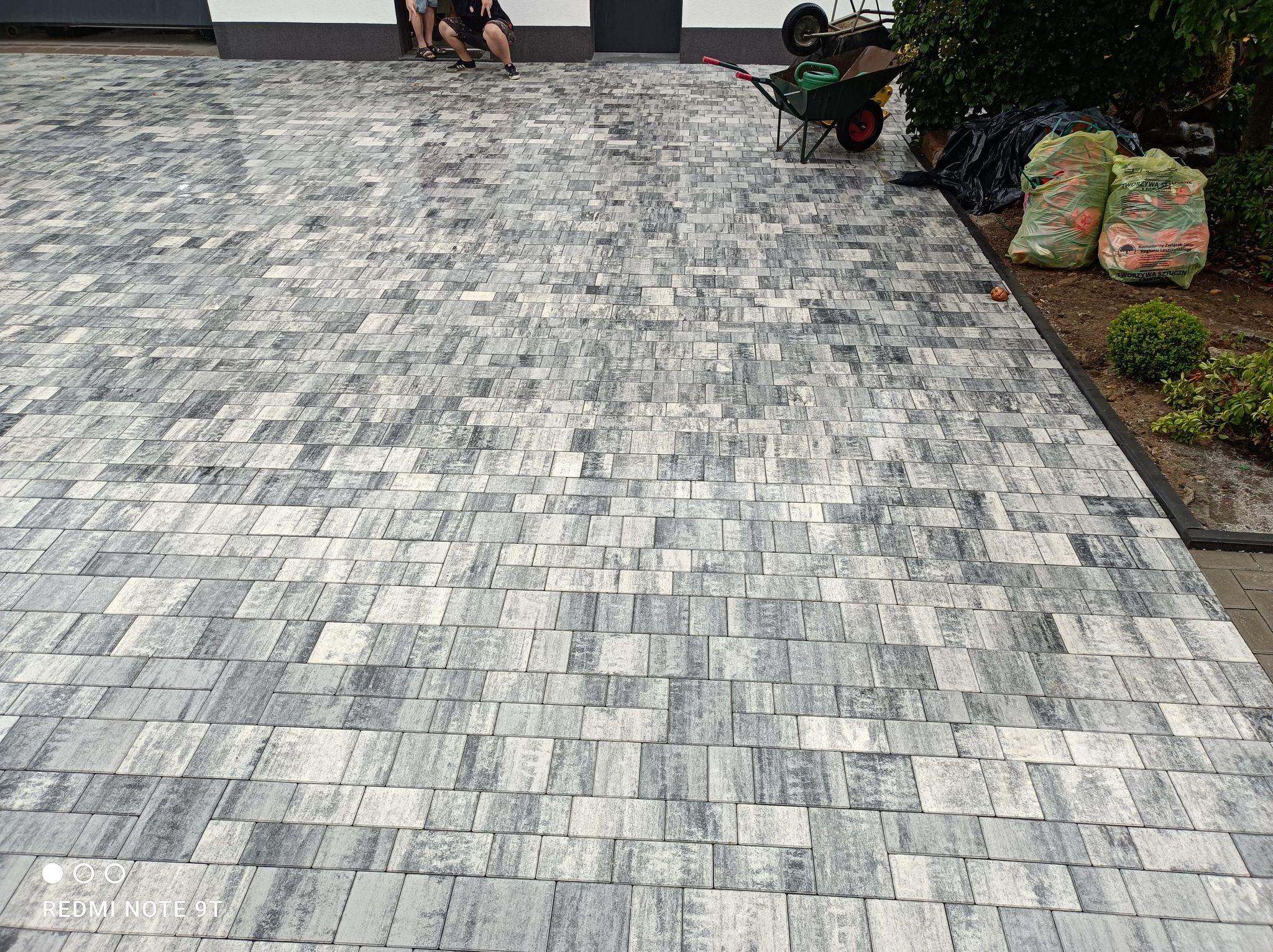 Ułożona szara kostka brukowa na posesji, widoczne worki z ziemią i taczka w tle.