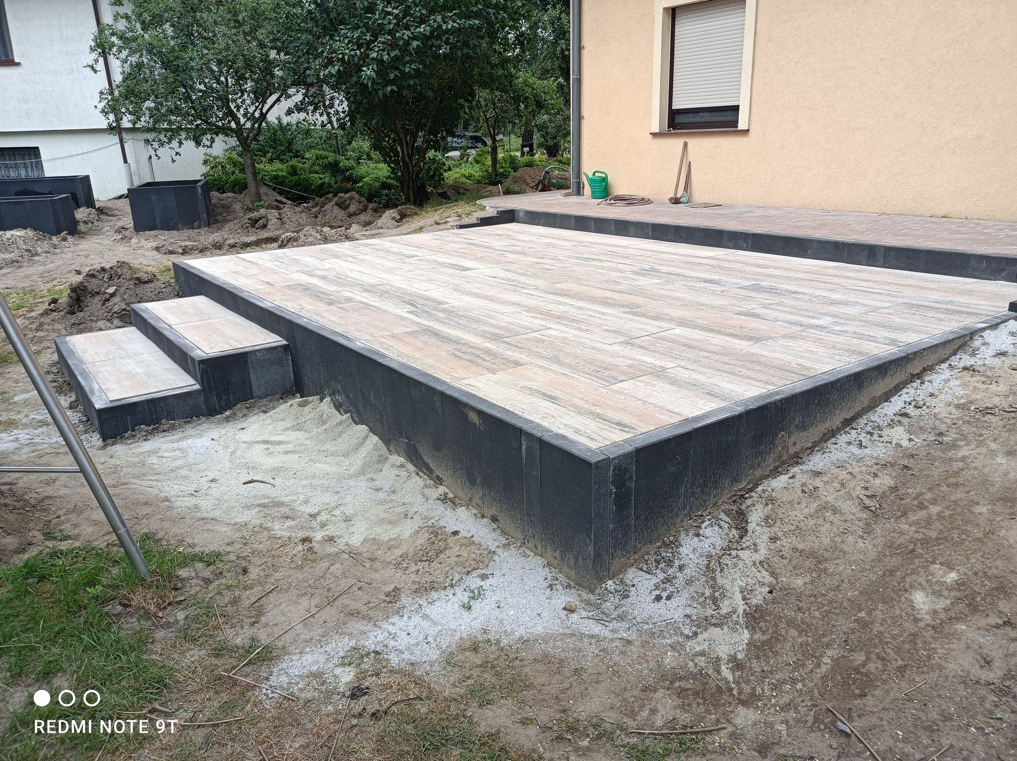 Wykończony taras z płytek imitujących drewno, obrzeża z ciemnego kamienia, widoczne schodki i przygotowane podłoże pod rabaty, dom z beżową elewacją w tle.