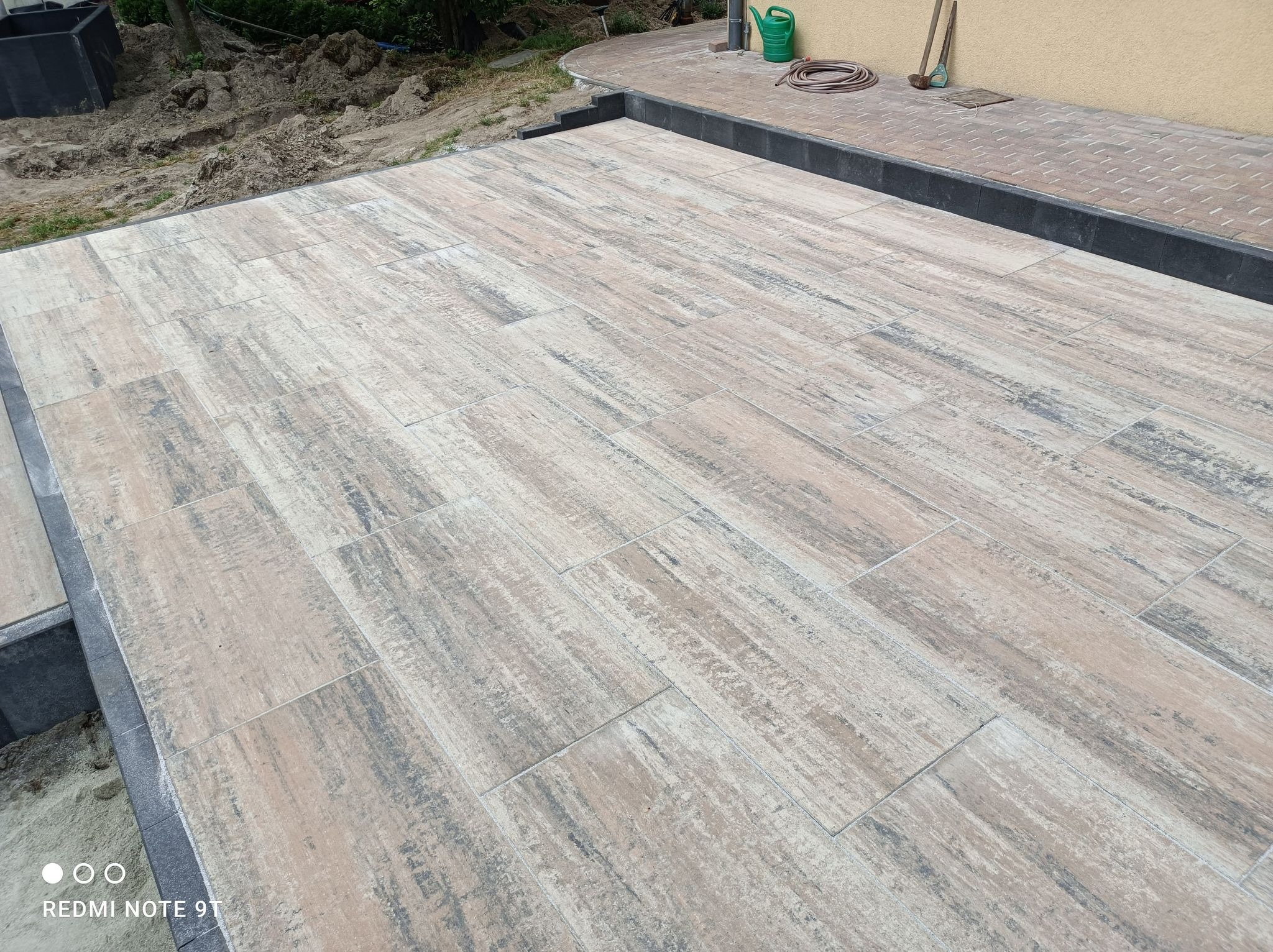 Ułożona kostka brukowa imitująca drewno na tarasie przydomowym, widoczne obrzeża z ciemnej kostki, w tle ziemia, trawa, konewka i wąż ogrodowy.