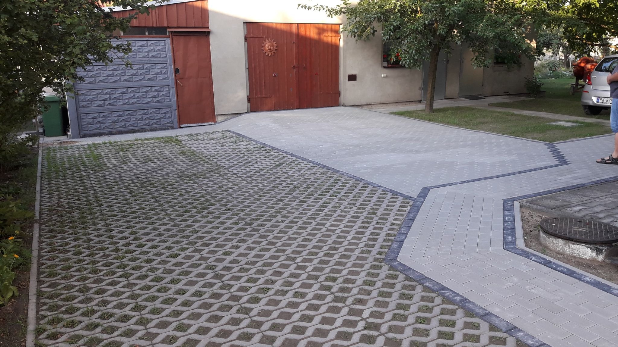 Ułożona szara kostka brukowa typu ażur z trawą wyrastającą w otworach, sąsiadująca z gładką kostką brukową z ciemnymi obramowaniami, prowadząca do garażu z drewnianymi drzwiami z ornamentem słońca.