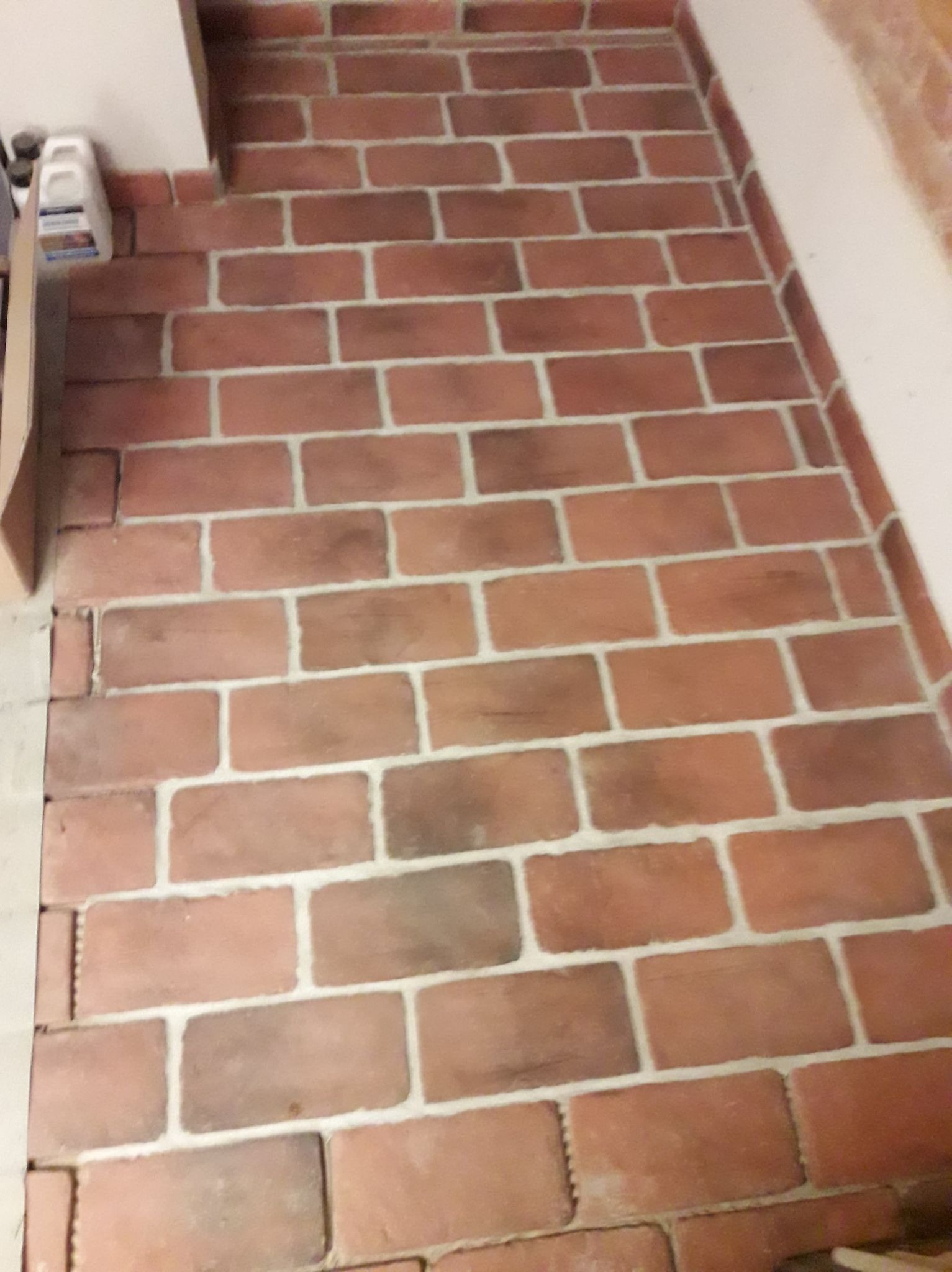 Ułożona ceglana podłoga z jasną fugą, widoczne przycięte cegły przy ścianie, butelka z płynem czyszczącym w rogu.