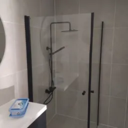 Nowoczesna kabina prysznicowa z czarnymi profilami i deszczownicą, jasne płytki na ścianach, widoczna umywalka z kosmetykami w tle.