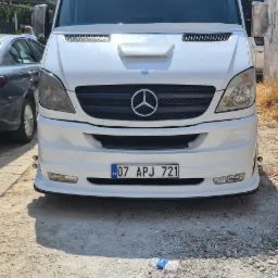 Biały, zmodyfikowany bus Mercedes-Benz Sprinter z naklejką 'DEMIROGLU TURIZM & TASIMACILIK' na przedniej szybie, zaparkowany na żwirowym podłożu.