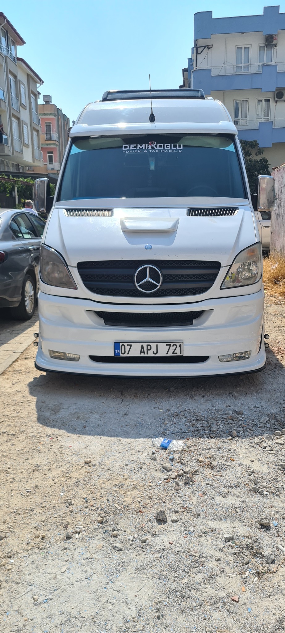 Biały, zmodyfikowany bus Mercedes-Benz Sprinter z naklejką 'DEMIROGLU TURIZM & TASIMACILIK' na przedniej szybie, zaparkowany na żwirowym podłożu.