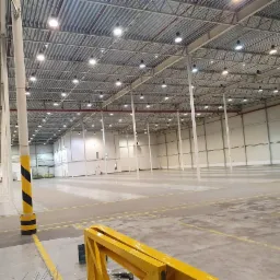 Wnętrze pustej hali magazynowej z metalową konstrukcją dachu i licznymi lampami sufitowymi, widoczne żółto-czarne oznaczenia na słupie i podłodze, w tle białe ściany.