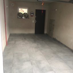 MATT WYKOŃCZENIA Mateusz Wrzosek - Wykończone wnętrze garażu z szarymi płytkami podłogowymi, widoczne dwie felgi samochodowe na parapecie okna, oraz fragment instalacji pod sufitem.