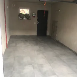 Wykończone wnętrze garażu z szarymi płytkami podłogowymi, widoczne dwie felgi samochodowe na parapecie okna, oraz fragment instalacji pod sufitem.