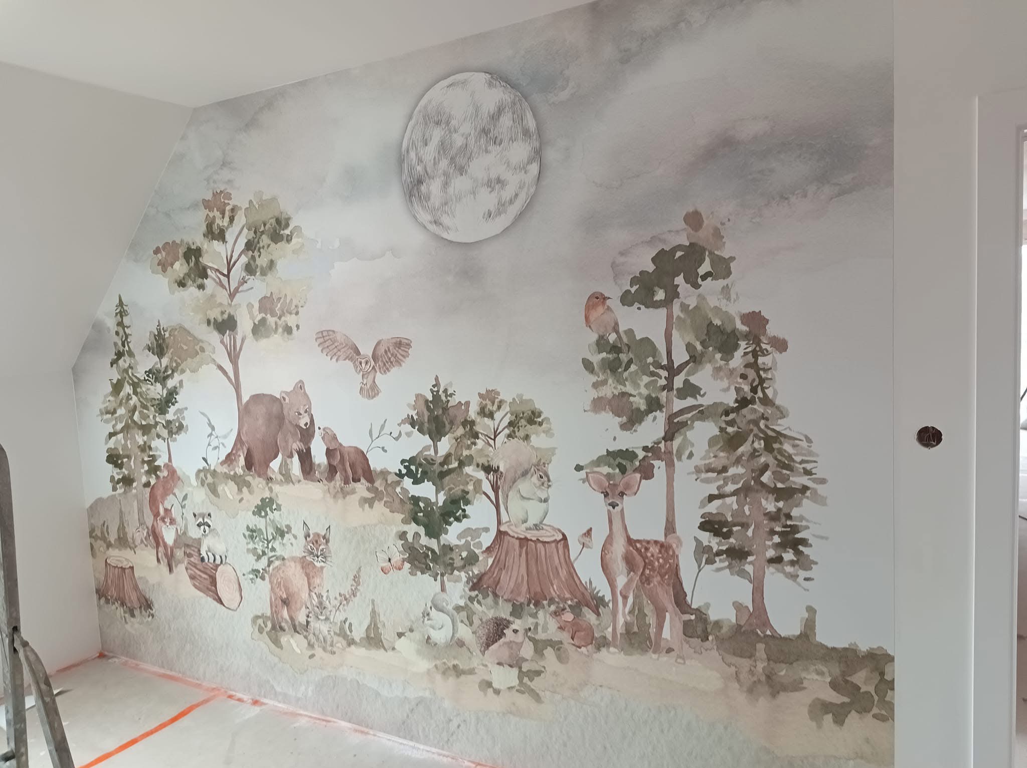 Pokój dziecięcy z ręcznie malowaną akwarelą na ścianie przedstawiającą leśne zwierzęta pod księżycem, widoczne niedoskonałości malowania przy podłodze.