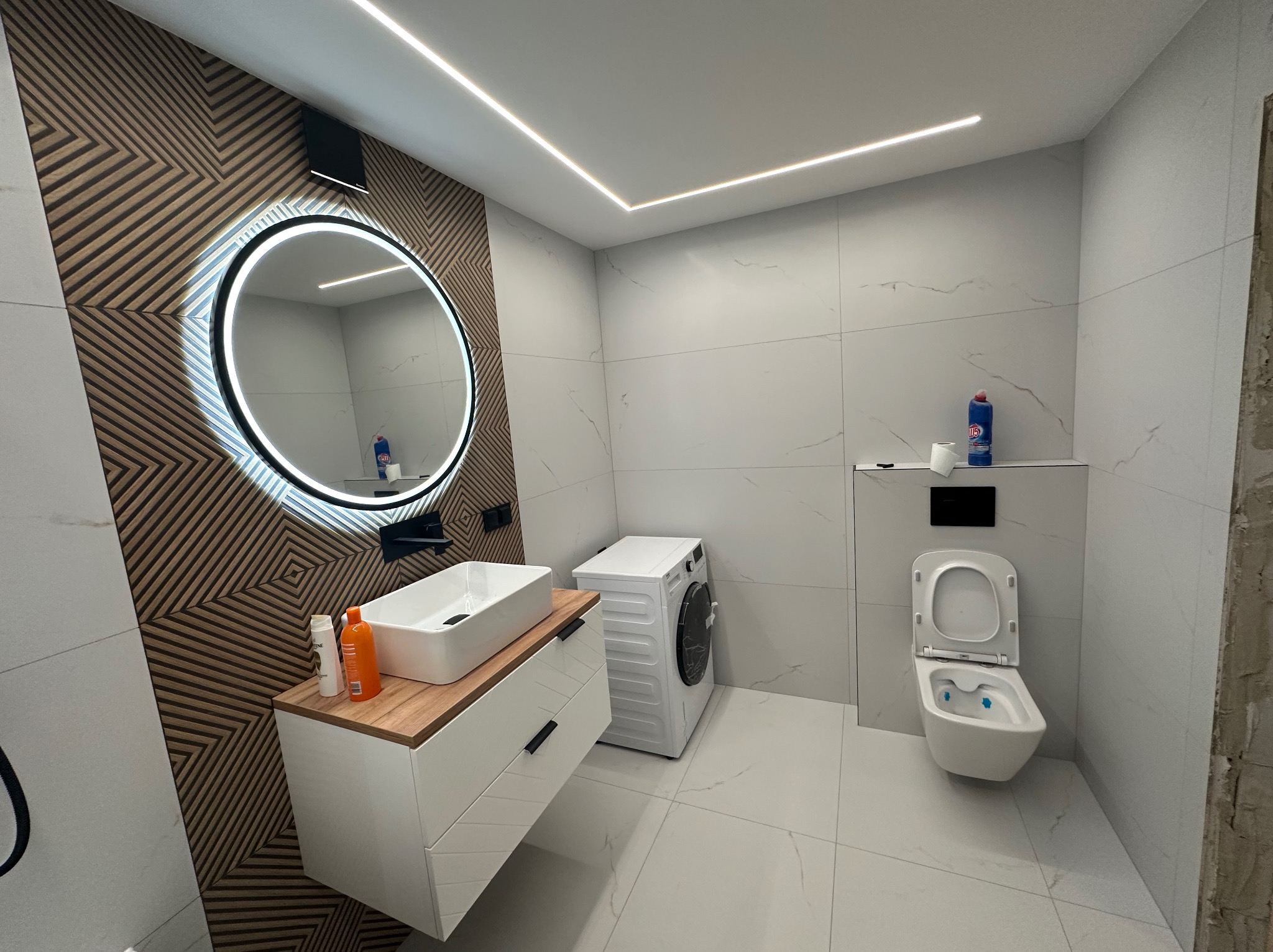 Nowoczesna łazienka z umywalką nablatową, okrągłym lustrem z podświetleniem LED, pralką i toaletą podwieszaną, wykończona białymi płytkami imitującymi marmur i drewnianą mozaiką na ścianie.