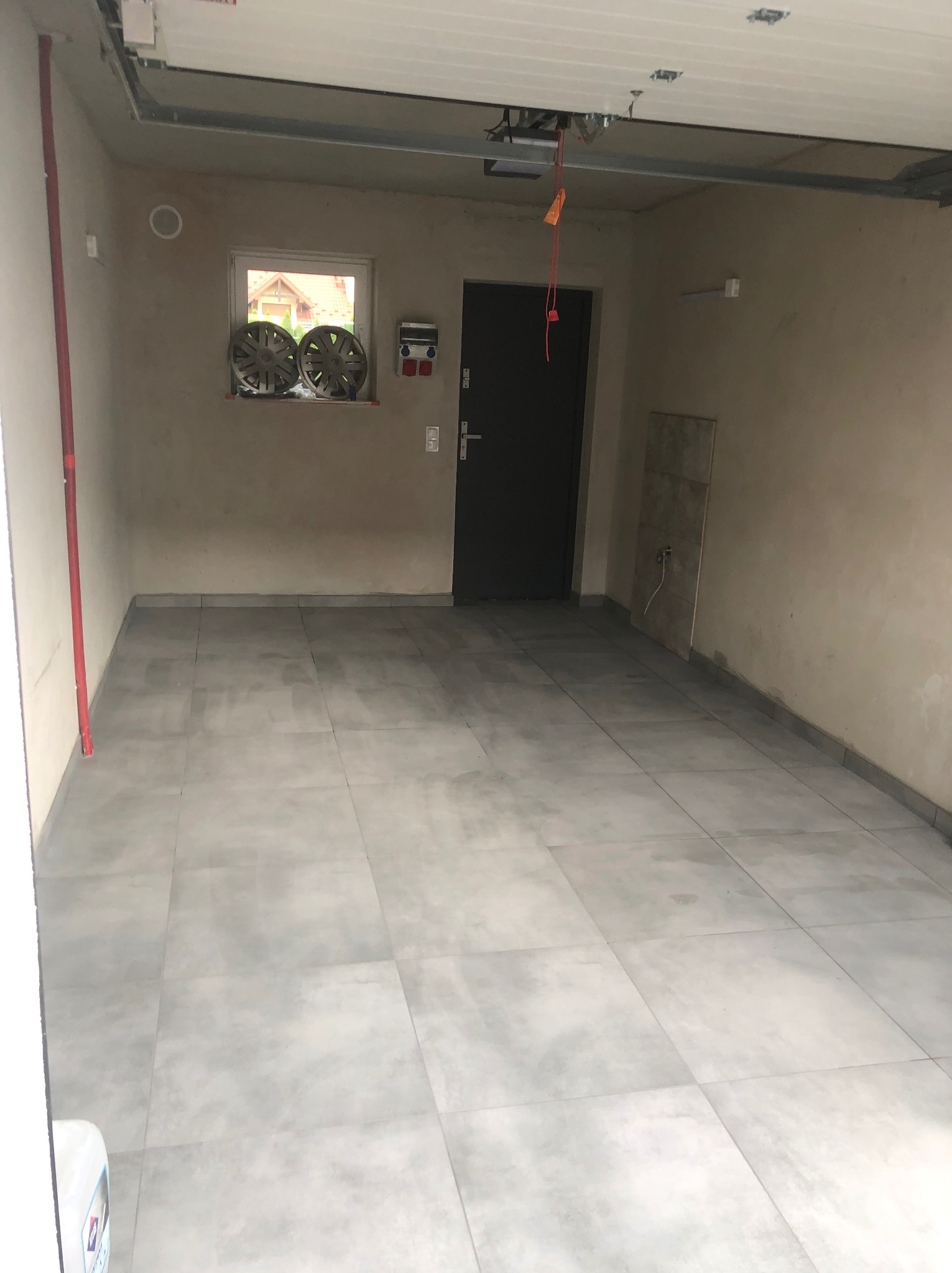 Wykończone wnętrze garażu z szarymi płytkami podłogowymi, widoczne dwie felgi samochodowe na parapecie okna, oraz fragment instalacji pod sufitem.