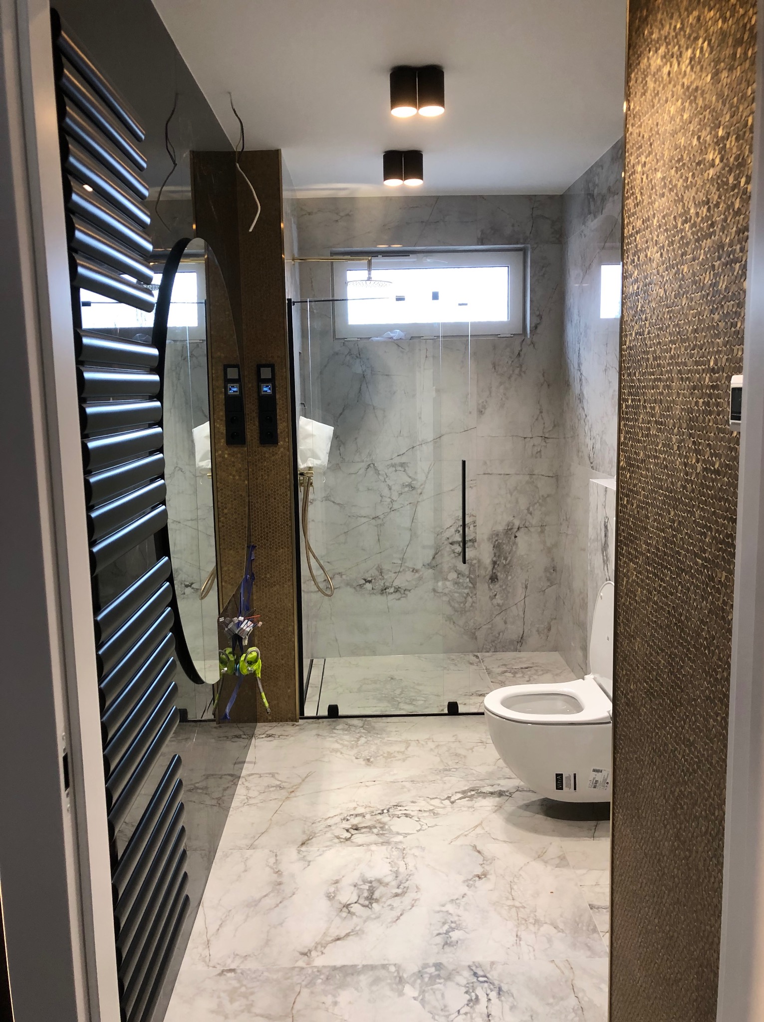 Nowoczesna łazienka z marmurową podłogą i ścianami, kabina prysznicowa ze szklanymi drzwiami, wisząca toaleta i grzejnik drabinkowy, widziana zza otwartych drzwi.