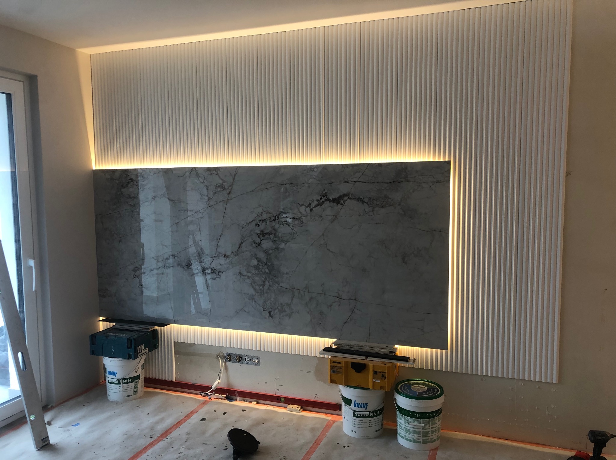 Ściana po remoncie z marmurową płytą i dekoracyjnymi, podświetlanymi listwami. Widoczne wiaderka z gładzią i narzędzia malarskie.