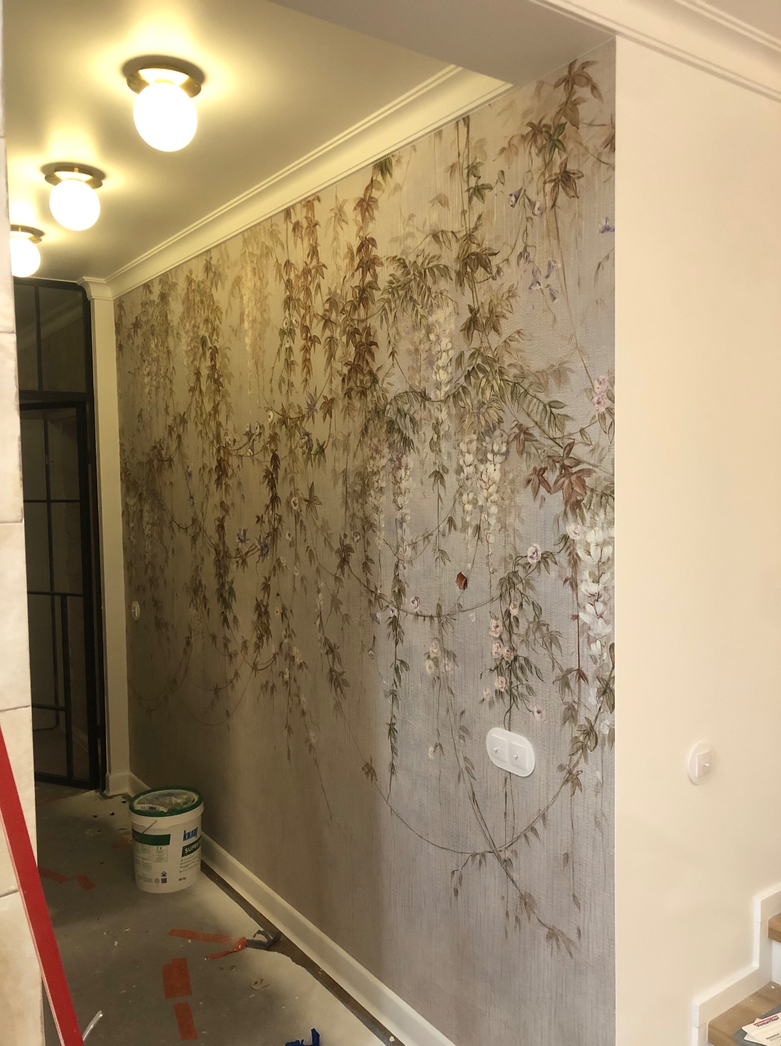 Korytarz w trakcie prac wykończeniowych z tapetą w motywy roślinne, widoczny kubeł z farbą i narzędzia na podłodze, oświetlenie sufitowe.