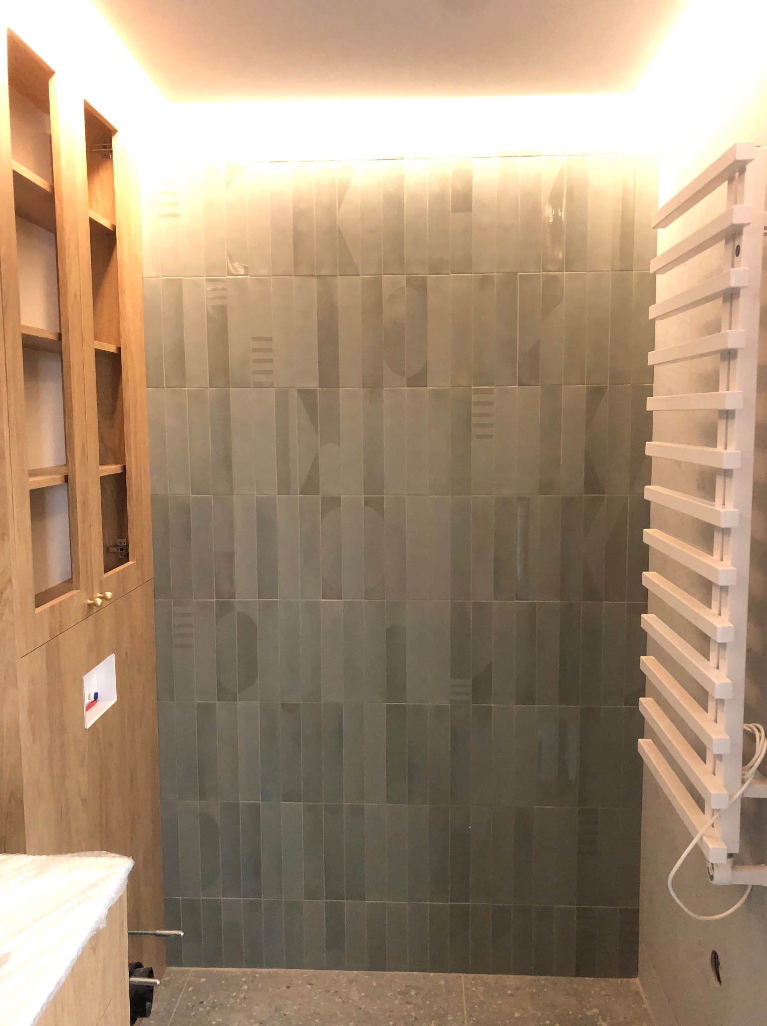 Wnętrze łazienki w trakcie remontu: pionowe, oliwkowe płytki z subtelnym wzorem, drewniana szafka z otwartymi półkami, biały grzejnik drabinkowy, szary betonowy podkład podłogowy, oświetlenie...