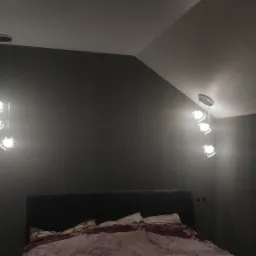 Sypialnia z ciemnoszarymi ścianami i dwoma wiszącymi lampami z trzema kwadratowymi kloszami, widoczne puszki elektryczne na ścianie.