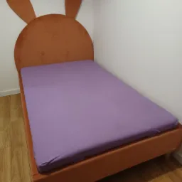 Łóżko tapicerowane w kolorze pomarańczowym z wezgłowiem w kształcie króliczych uszu, przykryte fioletowym prześcieradłem na drewnianej podłodze.
