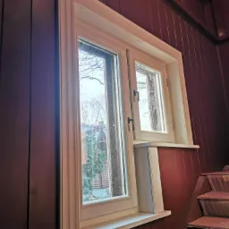 Białe, dwuczęściowe okno z widokiem na dom i drzewa, zamontowane w brązowej, drewnianej ścianie z pionowymi panelami, obok schody w trakcie remontu.