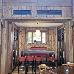 Drewniane wnętrze baru z wysokimi krzesłami, stołem i oknem z widokiem na zewnątrz, uzupełnione winiarkami i detalami snycerskimi.