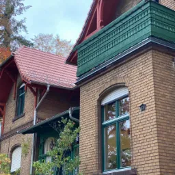 Dwupiętrowy budynek z cegły klinkierowej z czerwonym dachem i zielonymi elementami stolarki okiennej i balkonowej, widoczne rynny i numer budynku 33.