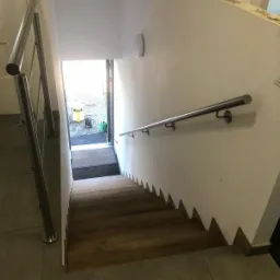 Schody drewniane z metalową poręczą, widok z góry, jasne wnętrze z wyjściem na zewnątrz.