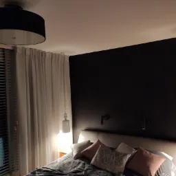 Sypialnia z czarną ścianą, łóżkiem z szarą pościelą i dekoracyjnymi poduszkami, oświetlona lampami wiszącymi i okrągłym plafonem, widok na okno z żaluzjami.