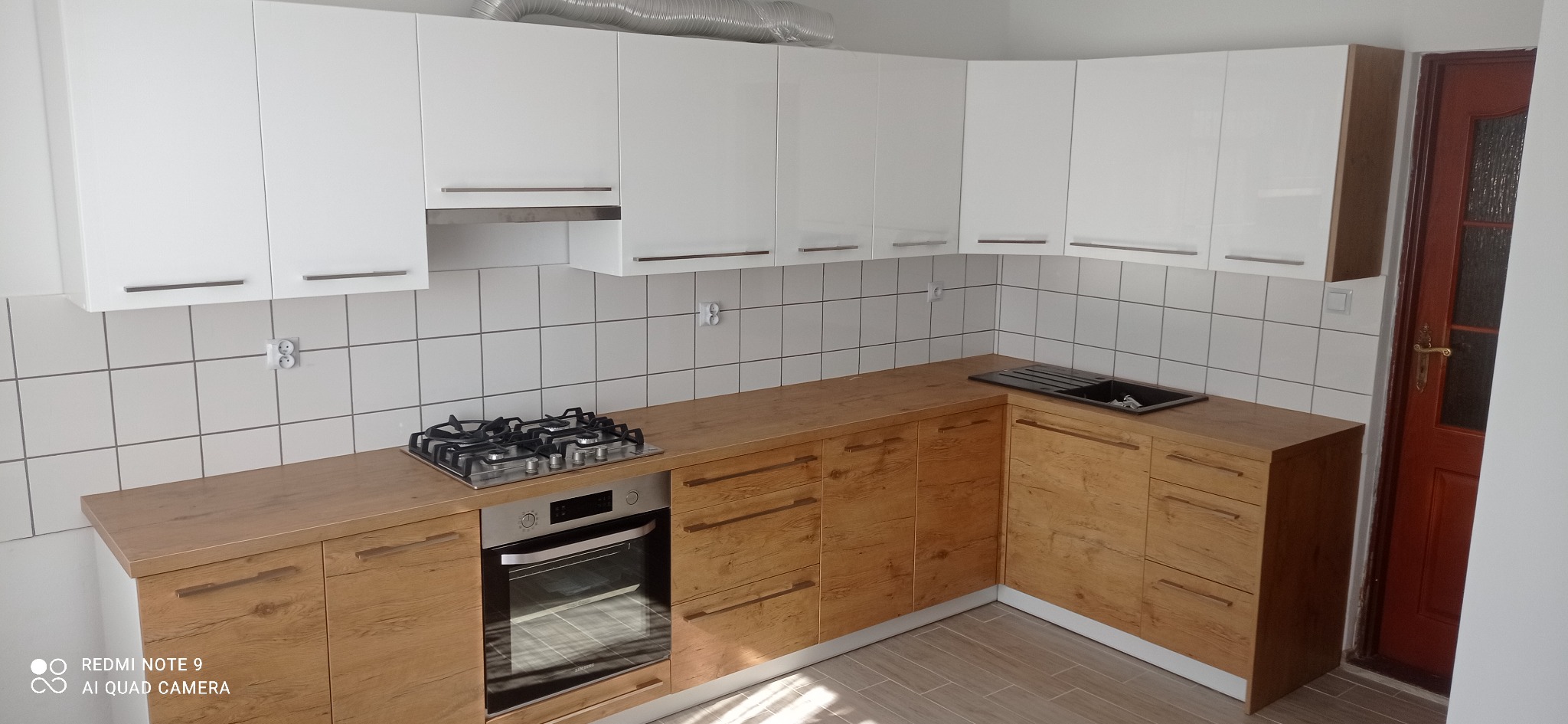 Nowoczesna kuchnia z białymi szafkami górnymi i drewnopodobnymi szafkami dolnymi, z widoczną płytą gazową i zlewem, na tle białych płytek.