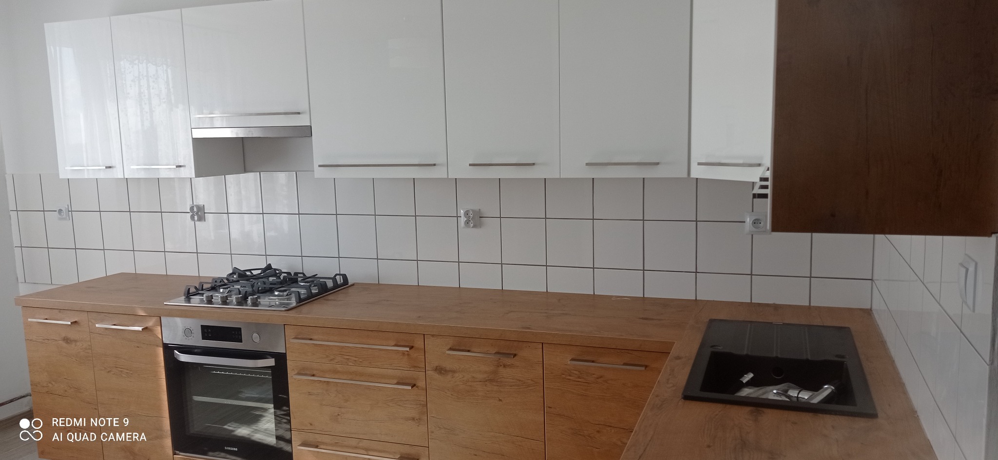 Nowoczesna kuchnia z białymi szafkami wiszącymi i drewnianymi szafkami dolnymi, widoczna płyta gazowa i czarny zlew.