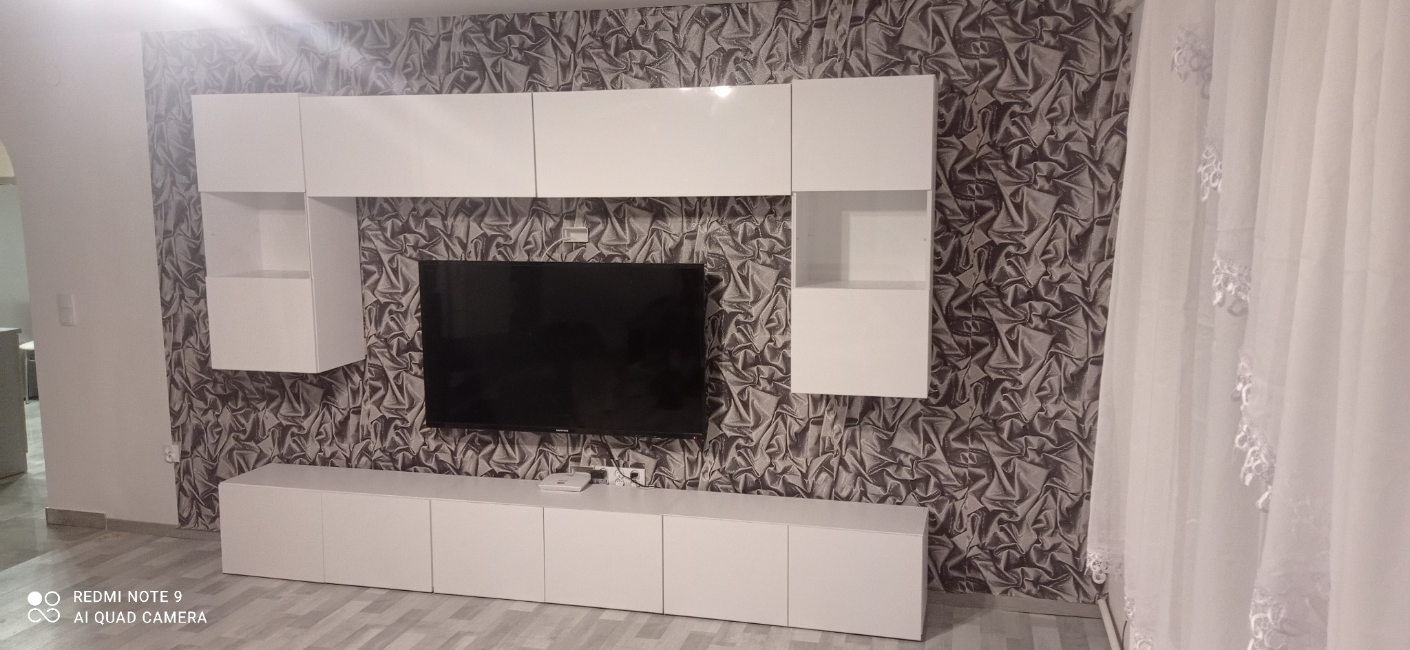 Biała, nowoczesna szafka RTV z wiszącymi półkami na tle wzorzystej tapety w salonie, z widocznymi przewodami i sprzętem elektronicznym.