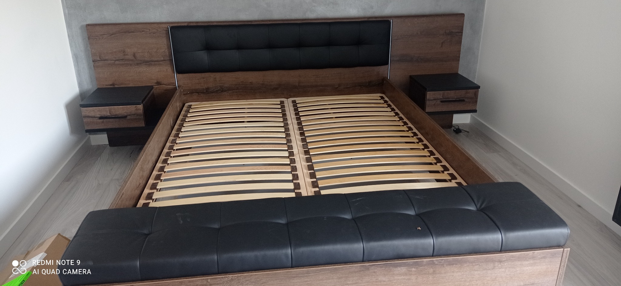 Drewniane łóżko z czarnym zagłówkiem i dwoma szafkami nocnymi bez materaca i pościeli, widoczny stelaż z listew