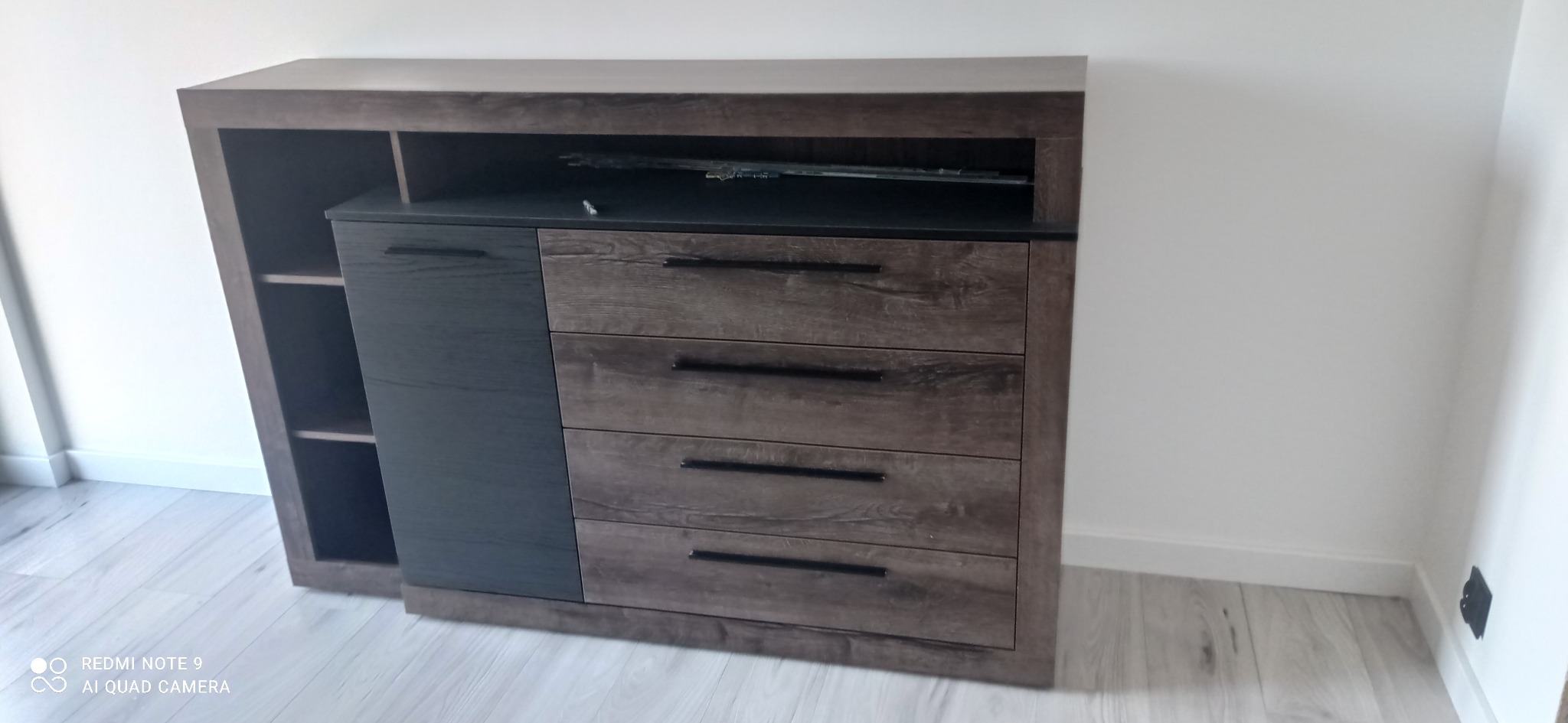 Nowoczesna komoda z szufladami i półkami, w odcieniach brązu i czerni, ustawiona przy białej ścianie na jasnej podłodze.