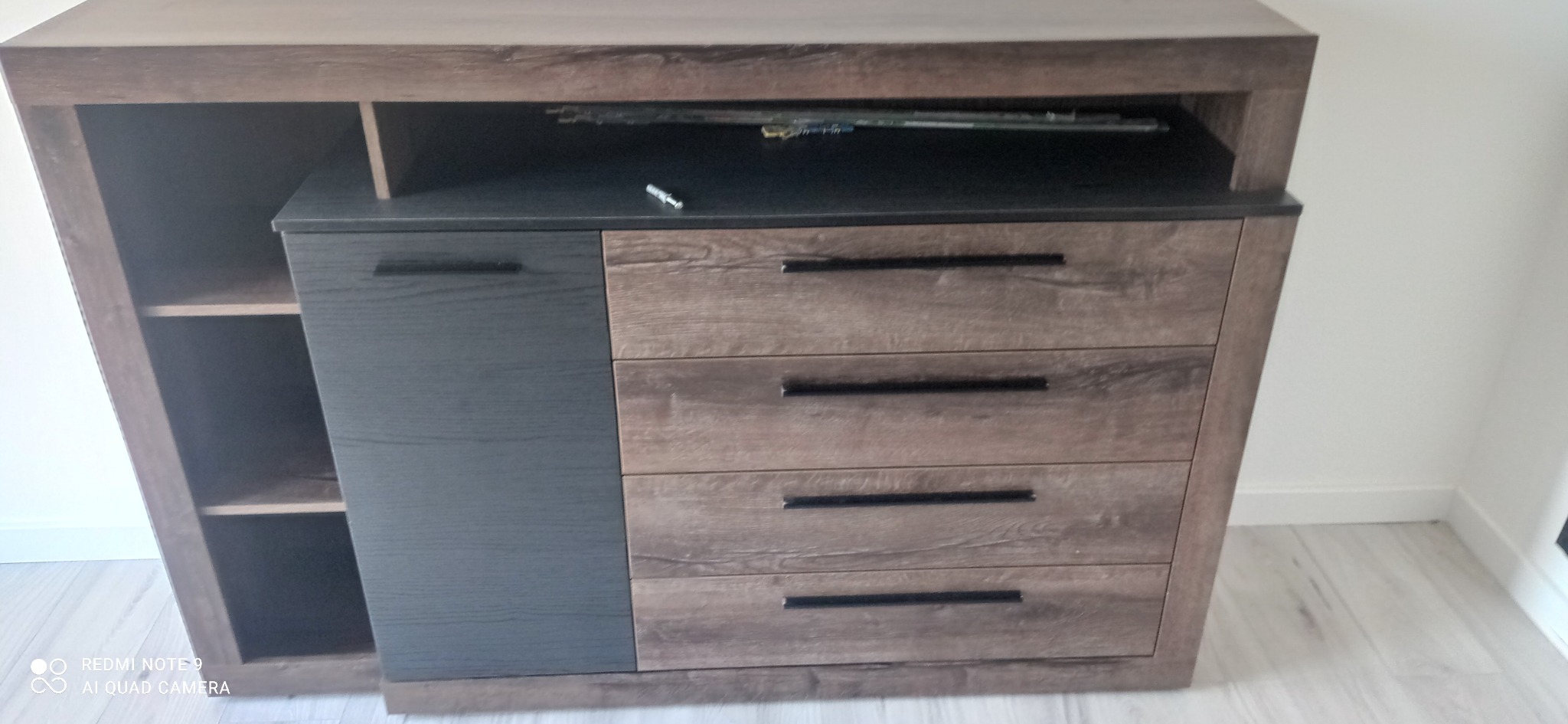 Komoda z szufladami i półkami, w odcieniach brązu i szarości, zamontowana w Tomaszowie Lubelskim. Na blacie komody leżą klucze.