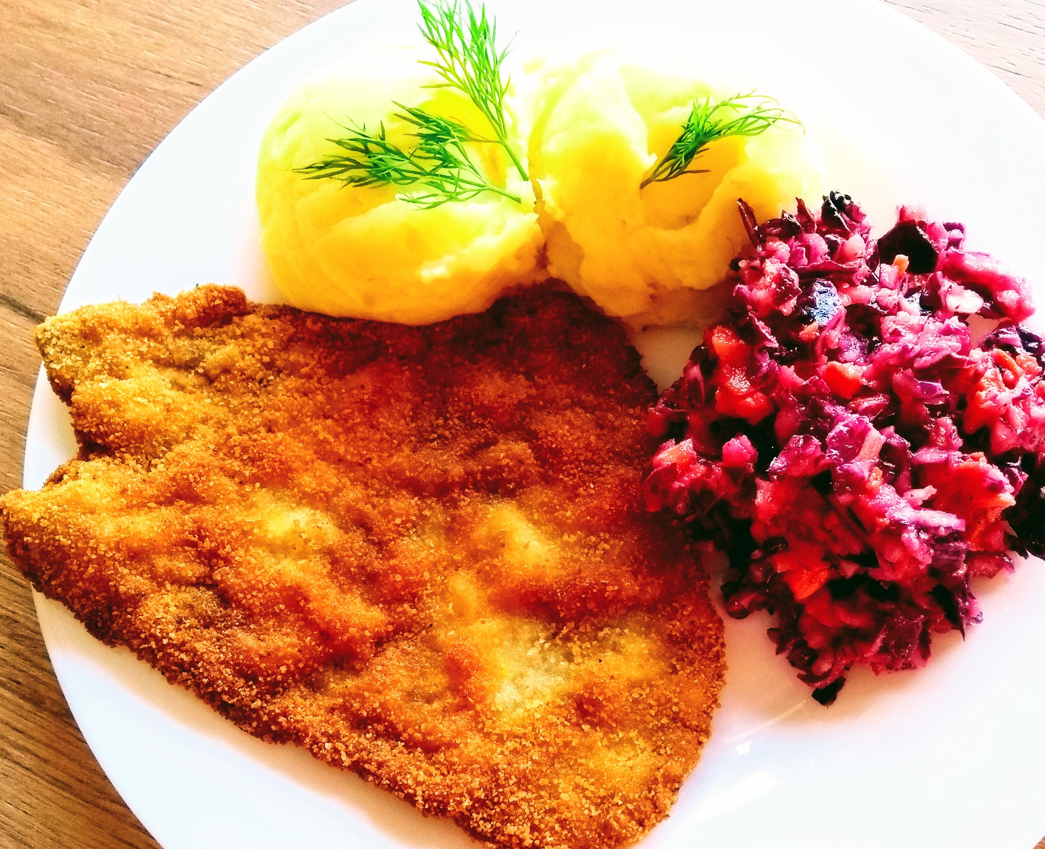 Kotlet schabowy w złocistej panierce z ziemniaczanym puree dekorowanym koperkiem i surówką z czerwonej kapusty na białym talerzu.