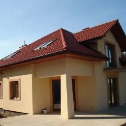 Wykończony dom jednorodzinny z beżową elewacją, czerwoną dachówką i oknami dachowymi, widziany z perspektywy ogrodu w słoneczny dzień.