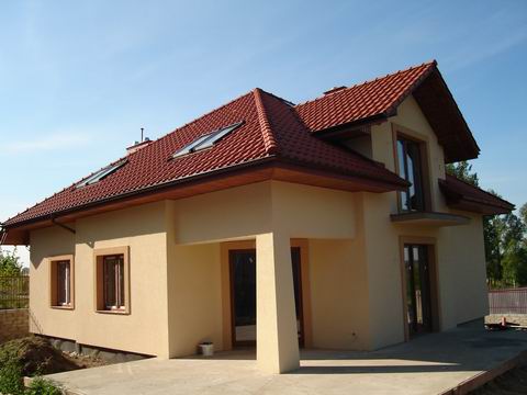 Wykończony dom jednorodzinny z beżową elewacją, czerwoną dachówką i oknami dachowymi, widziany z perspektywy ogrodu w słoneczny dzień.