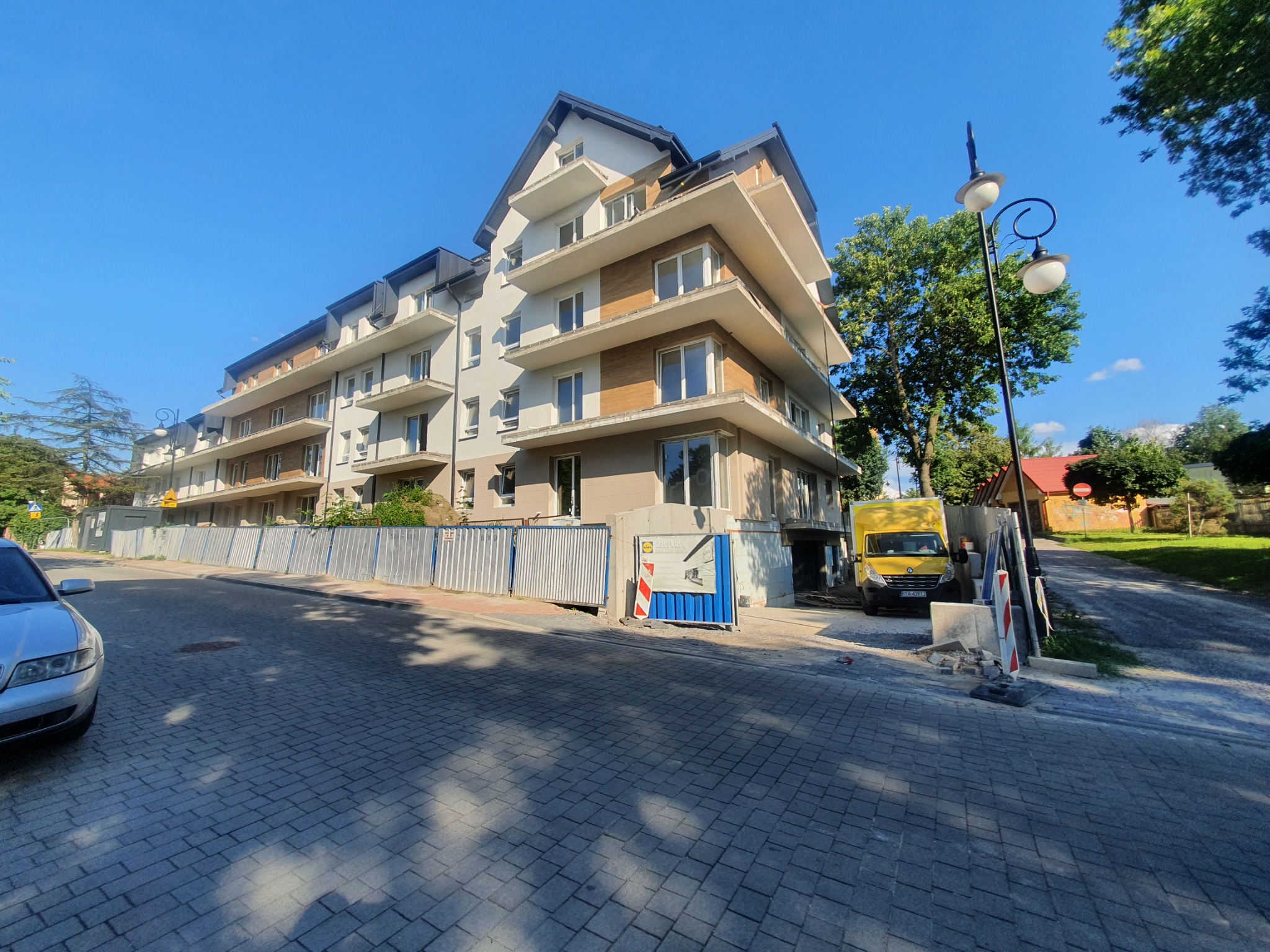 Wielopiętrowy budynek mieszkalny w trakcie wykańczania elewacji, widoczny z poziomu ulicy, z zaparkowanym żółtym samochodem dostawczym i elementami zabezpieczeń budowy.