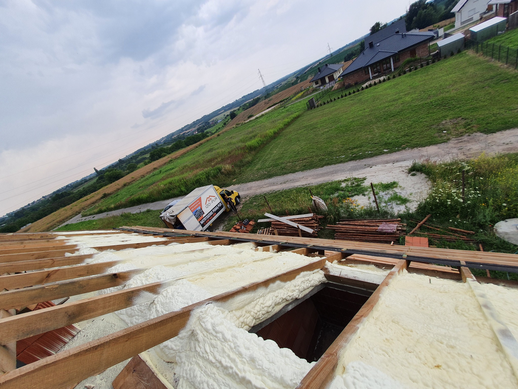 Poddasze w trakcie ocieplania pianką PUR, widoczne drewniane krokwie, biała pianka izolacyjna, na zewnątrz krajobraz z domami i samochód dostawczy z logo firmy IZO-PUR.