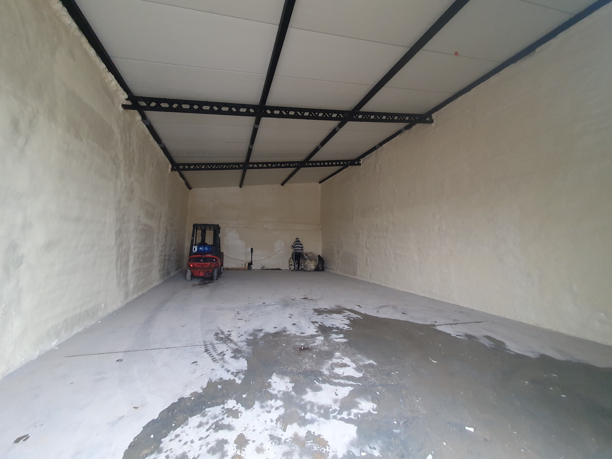 Wnętrze hali przemysłowej po ociepleniu pianką PUR, widoczny wózek widłowy i pracownik przy workach, mokra posadzka betonowa.