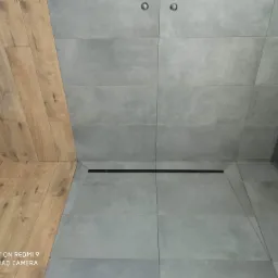 Szare, matowe płytki w kabinie prysznicowej z odpływem liniowym, kontrastujące z drewnopodobną okładziną ściany, widok z góry.