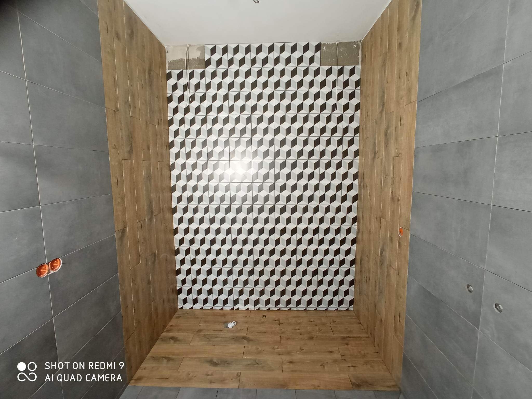 Wnętrze łazienki w trakcie wykańczania: ściana z płytek w geometryczny wzór 3D, panele drewnopodobne na ścianach bocznych, szare płytki na pozostałych ścianach, podłoga wyłożona panelami...