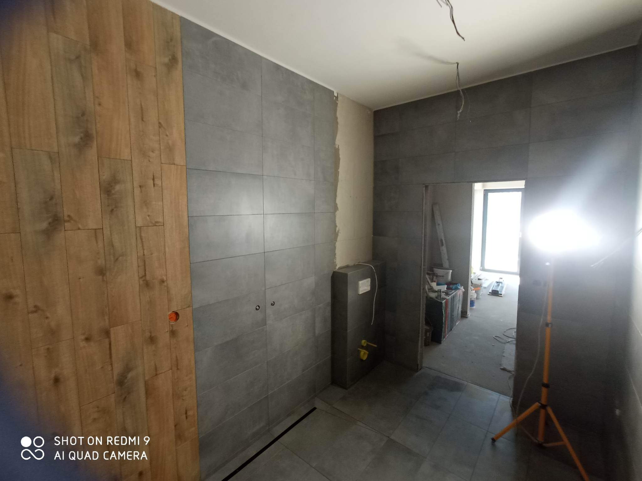 Układanie szarych płytek w łazience w Poznaniu, z fragmentem ściany wykończonej panelami drewnopodobnymi. Widoczne przygotowania do montażu armatury i oświetlenia.