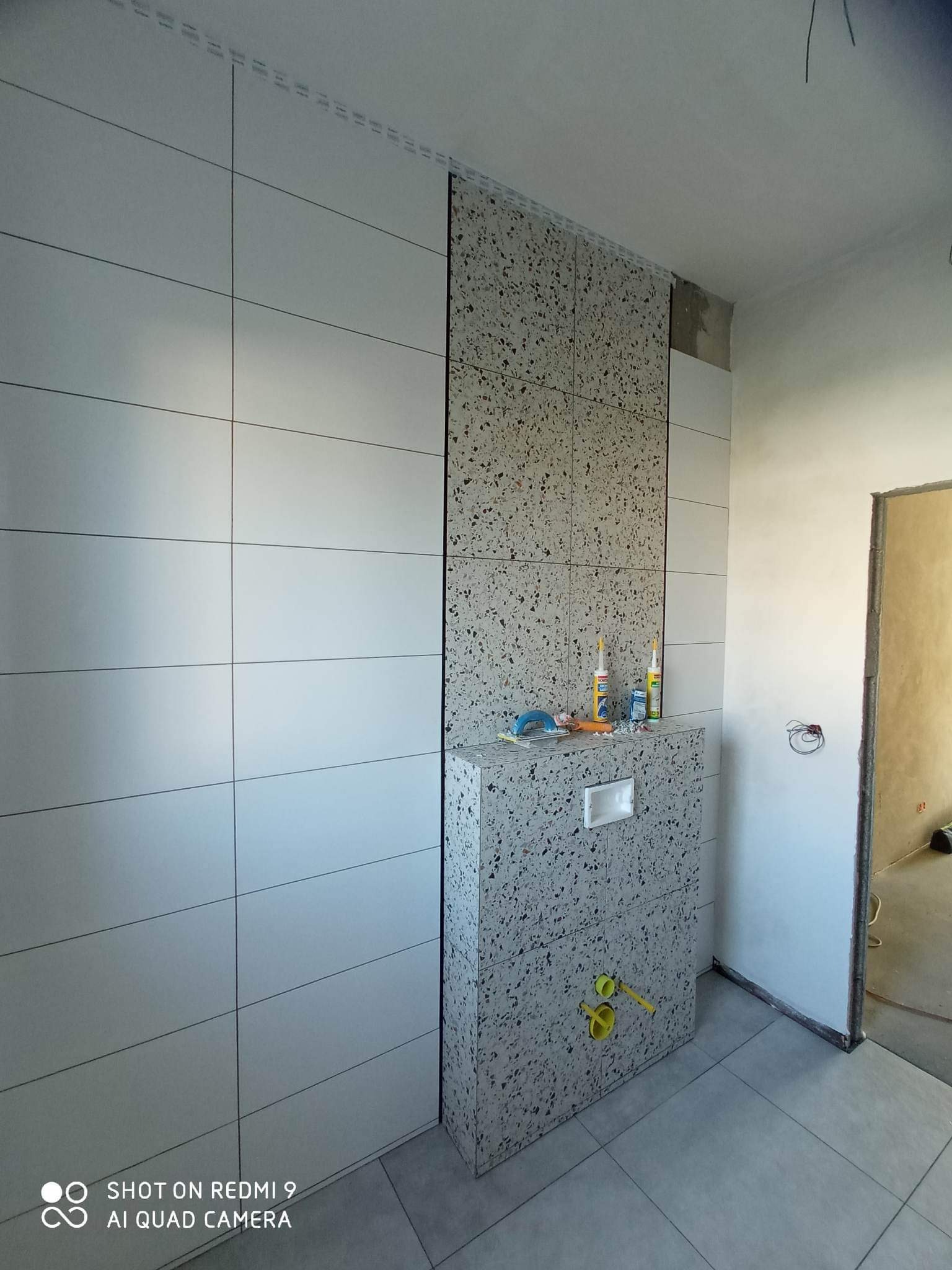 Ściana łazienki w trakcie wykańczania: białe, prostokątne płytki ceramiczne zestawione z panelami z mozaiki lastryko, widoczne narzędzia i materiały budowlane, instalacja sanitarna w trakcie montażu.