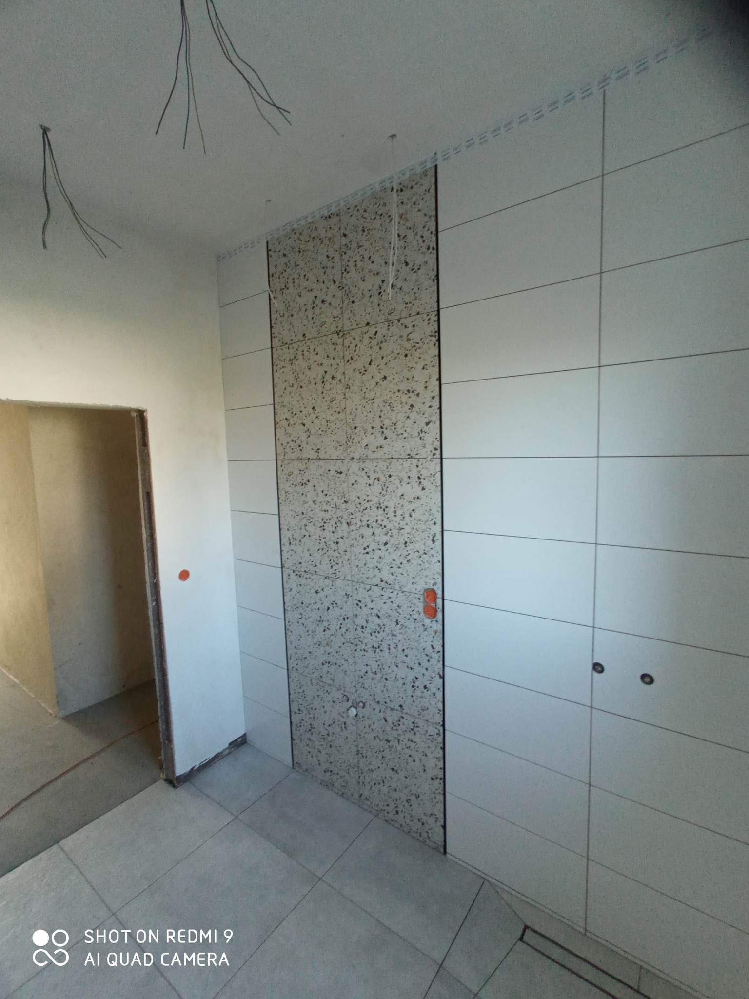 Świeżo wykafelkowana ściana z dwoma rodzajami płytek: gładkie, poziome białe płytki oraz pionowy pas z płytek imitujących lastryko. Widoczne przewody elektryczne wystające z sufitu i otwór drzwiowy...