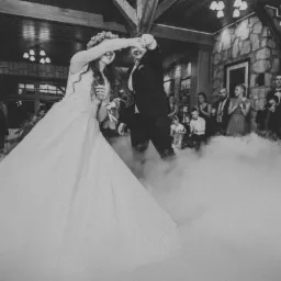 Czarno-białe zdjęcie pary młodej podczas pierwszego tańca w chmurze dymu na sali weselnej z kamiennymi ścianami, oświetlonej żyrandolami, w tle goście weselni.