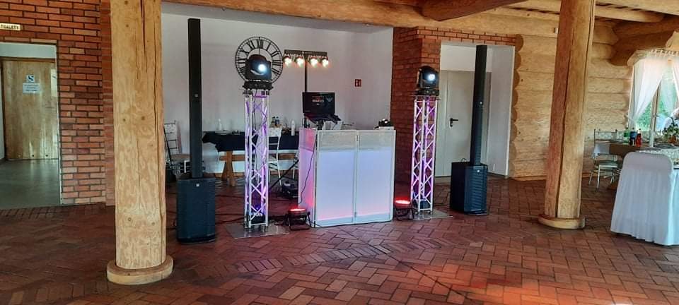 Sprzęt nagłośnieniowy i oświetleniowy, w tym kolumny głośnikowe, reflektory na statywach z motywem zegara i podświetlany panel DJ-a, ustawiony w rustykalnym wnętrzu z drewnianymi kolumnami...